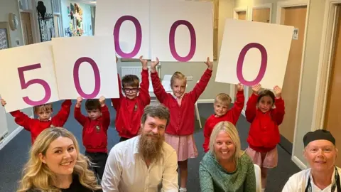 Burton Green Primary School children hold up individual signs with numbers to represent 50,000 school meals have been served as part of the Hungry Minds Appeal scheme. Staff members and councillor Bob Webb sit in front of the children. 