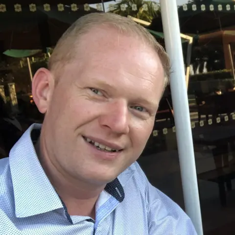 Tristan Campbell A man with short ginger hair smiling at the camera. He is wearing a blue shirt. 
