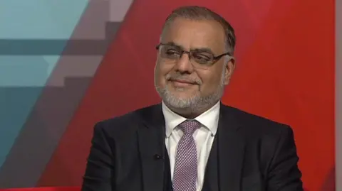 Independent MP Ayoub Khan. He is wearing a three-piece suit with a smart dotted tie. He has a beard and is wearing glasses.
