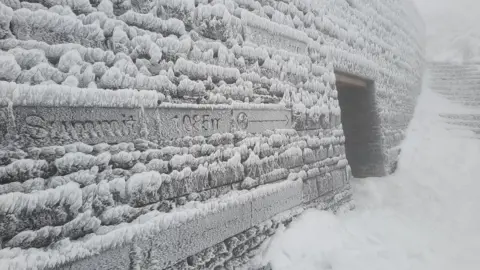 Tony Ellis Frost, ice and snow covers the walls and entrance to a building at the summit of Yr Wyddfa. A sign says 'summit'.