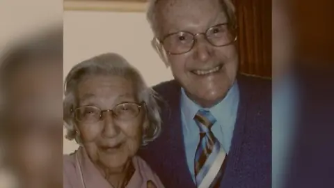 Family photo Head and shoulders portraint photo of Gladys and Ron, both smiling to the camera. Gladys has bobbed grey straight hair and Ron has short cropped grey hair. Both are wearing metal rimmed glasses.