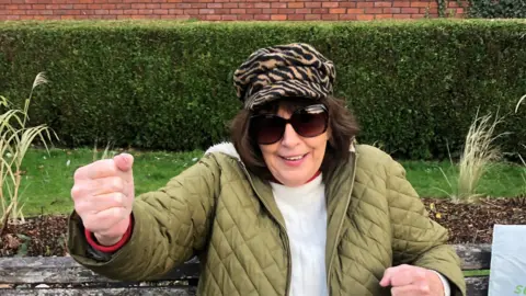 Lynda Debono is sat on a bench, she is wearing a white jumper and green jacket with an animal print hat and oversize sunglasses.