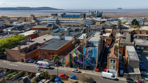 A view over Weston with the beach in the background. There is some colourful artwork on some of the house walls in the foreground of the picture.