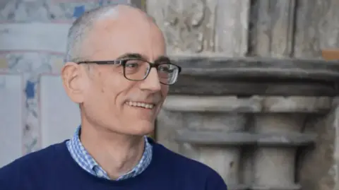 Lincoln Cathedral Jeremy Wheeldon, a man with short grey hair and glasses, is smiling. He is wearing a blue and white check short under a navy jumper.