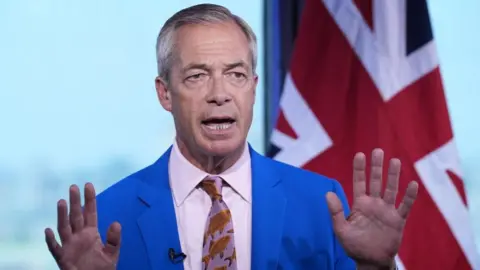 Nigel Farage is wearing a bright blue coloured suit jacket, white shirt and patterned tie. He is stood in front of a Union flag. His hands are held up in front of him.