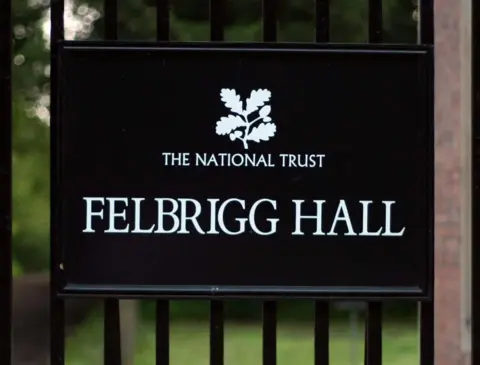 PA Media A black rectangular sign mounted on metal gate railings features a white oak leaf logo and "THE NATIONAL TRUST" at the top, with the words "FELBRIGG HALL" written below it in a large, clean, white serif font.