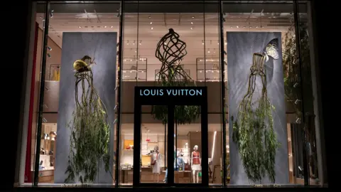 Full Grown Two chairs grown from trees hang in the windows of a Louis Vuitton store in London in 2022. The chairs are lifted up with branches and leaves dangling from the legs of the chairs. In the middle of the store window is a chandelier also grown from a tree.