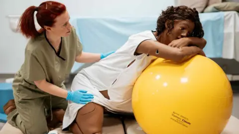 Rebecca Hudson-Tandy, lecturer in midwifery touches the back of Mama Anne, a human-like manikin. Mama Anne resembles a pregnant person, wearing a white hospital gown. It leans over a large ball, head in hands. 