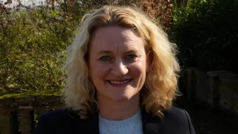 John Fairhall/BBC Louise McKinlay has curly, shoulder-length blonde hair. She is wearing a dark jacket over a baby blue jumper. She is smiling in front of bushes outdoors.