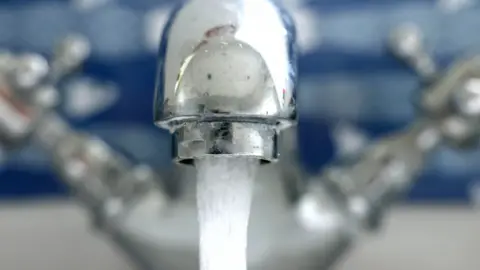 The picture shows a close-up of a chrome bathroom or kitchen tap with water running from it. The tap has two handles, one on each side. The water flow appears steady and clear, suggesting clean running water. The background is slightly blurred but seems to feature a blue and white patterned surface, possibly tiles or a decorative panel. 
