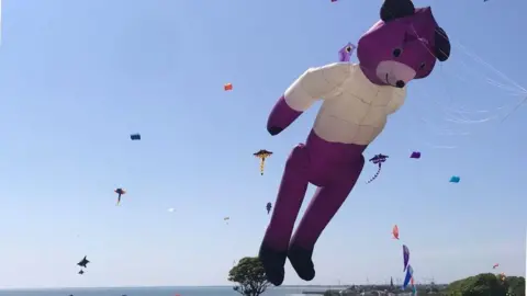 A kite in the shape of a large purple bear flies at the Bridlington Kite Festival. Other colourful kites are visible in the background and are set against a blue, sunny sky.