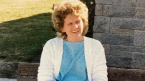 Family handout A slightly faded, older photograph taken outdoors in bright sunlight. In the centre foreground, Joan is sitting on a wooden bench. She is wearing a light blue dress and a white cardigan, with her legs crossed and her hands resting in her lap, holding a small dark handbag with a thin strap.