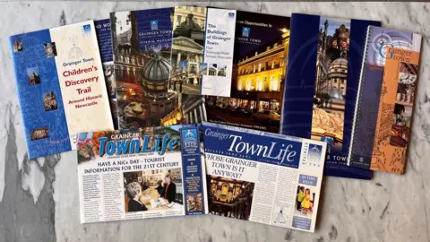 Newcastle City Council Magazines folded over which read 'Grainger Town Life' and other colourful blue booklets lie on a table in a spread.