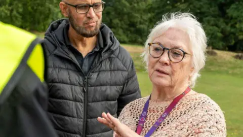 Northamptonshire OPFCC Danielle Stone, to the right, with her hand out, wearing a knitted top, with a lanyard round her neck. She has on glasses and has short fair hair. She is standing by a man wearing a black coat, with a black T-shirt, with glasses and a beard, he is looking at her. They are in a field with trees and grass behind her. 