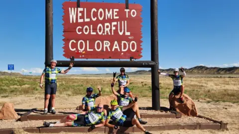Mike Teague A group of men in brightly coloured cycling outfits posing in front of a sign that reads 'Welcome to Colorado'