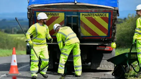 Three workmen in high-vis clothing are working behind a Highway Maintenance van. There is an orange cone to one side of the narrow countryside road. They are wearing white, hard hats and sturdy black boots. 