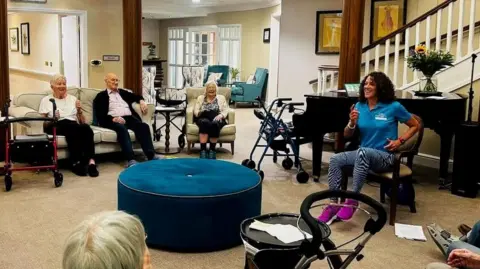 Jules Jenkinson Jules Jenkinson leads a group of care home residents in an exercise session. Every one is sat down in chairs in a circle in a wide open space in the home. 