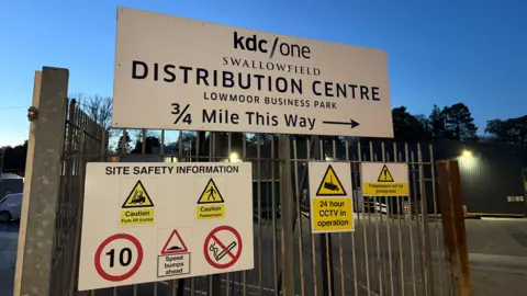 An entryway into the factory. There is a large sign on the gate which says 'KDC/One, swallowfield, distribution centre, lowmoor business park, 3/4 mile this way'. It is getting dark.