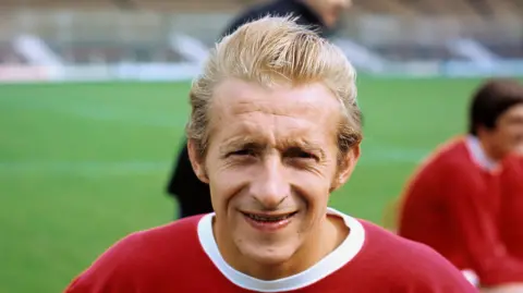 PA Media Denis Law on a football pitch, looking at the camera in his younger football days. He is smiling and wears a red top