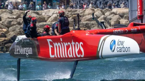 Great Britain's SailGP team celebrate on their catamaran after crossing the finish line. The craft has large sponsor logos for Emirates and JP Morgan.