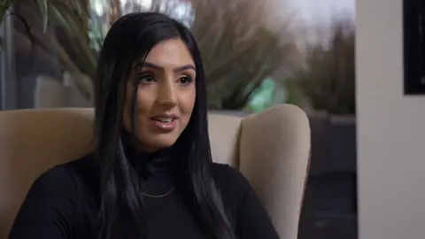 British Asian woman sitting on a cream sofa. She has black long hair and wearing a black turtle neck jumper with a gold thin chain around her neck. There are plants in the background.