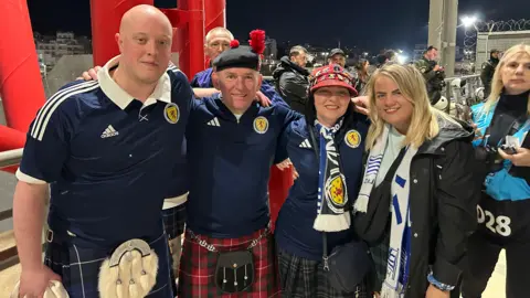 Thomas Mackintosh dressed in a kilt and navy blue Scotland football shirt, alongside three Scottish football fans in shorts, scarves and kilts, at a match