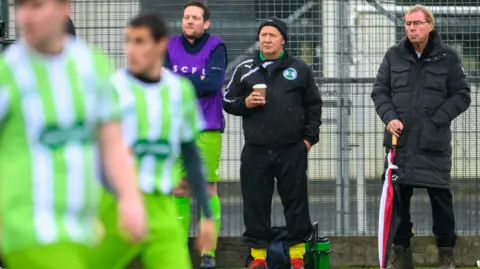 Tunley Athletic FC Harry Redknapp on the sidelines standing next to the permanent coach, with a couple of blurred footballers in the foreground in play