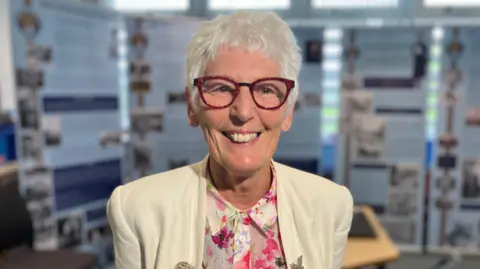 BBC/Sam Read A smiling Stephanie Daniels looks directly at the camera wearing large, red-rimmed glasses, a white jacket and blouse with a flowery print. 