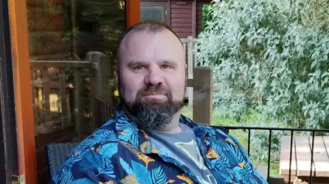 BoB Mark Willerton in his wheelchair on a balcony, wearing a summery short-sleeved blue shirt and grey shorts.
