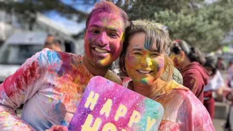 BBC A man and a woman covered in colourful powder smile and pose while holding a sign that says Happy Holi.
