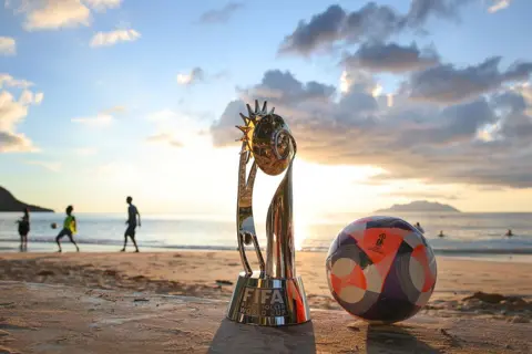 ALEX GRIMM / GETTY IMAGES A detailed view of the trophy and the official match ball as people play football at Beau Vallon beach. The sun is low is the sky above the sea in the background.
