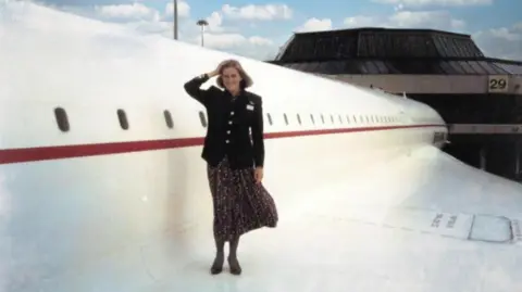 Julie Reynolds An old photo of Julie Reynolds stood on the wing of a Concorde jet. She is in cabin crew uniform and has one hand on her head. She is looking at the camera and smiling.