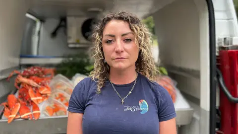 Gabby Mason has short, curly hair, she stands at the open back of a white van, wearing a navy T-shirt with a “Jade’s Fisheries” logo. Inside the van, trays and baskets filled with bright orange shellfish are visible, along with equipment and storage containers. The van’s interior frames the scene, with greenery faintly visible in the background.