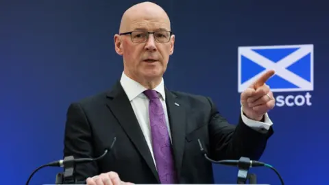 A bald man with glasses points with his left hand while speaking at a podium. He is wearing a dark suit, white shirt and purple tie. There is a blue background behind him with a saltire on it 