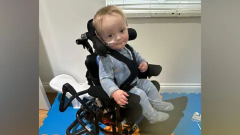 Rainbow Trust A young child in a wheelchair. He has a tube from his nose, indicating he is unwell. He is looking at the camera.
