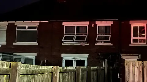 The house where the fire broke out. It is a terraced house surrounded by other homes. Three upstairs windows have been opened. A pink curtain is hanging out of the centre window.