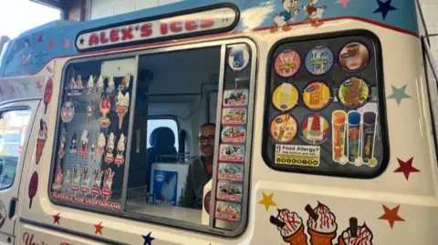 The side of an ice cream van, with pictures of ice creams and lollies in the windows of the vehicle. A man can be seen sat at the serving counter