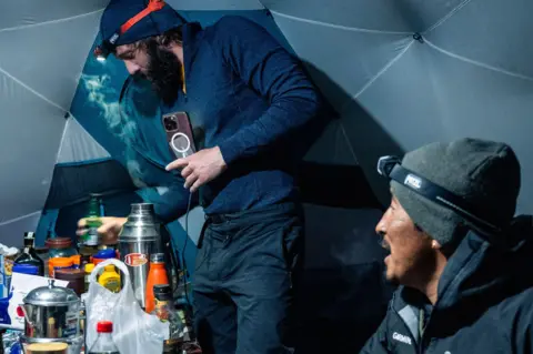 Blue Door Productions/Stan Gaskell Two men inside a tent. There is food on the table and lots of drink bottles. One of the men is Hutchcraft. The other is guide Gelje Sherpa.