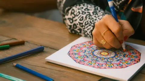 A person draws a colourful round pattern using pencils and pens. They are wearing a white and black animal print shirt and gold rings.