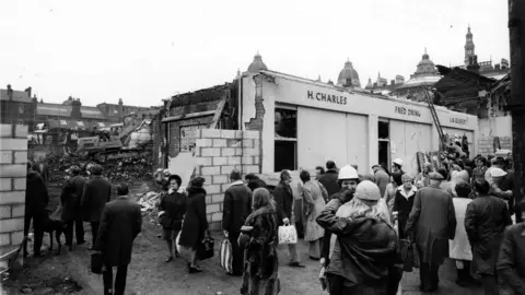 Leeds City Council A black-and-white photo showing a crowd gathered around a partially demolished building with visible signage reading “H. Charles,” “Fred Dining,” and “L. Gilbert.” The structure appears damaged, with broken walls and rubble scattered nearby. People in coats and hats stand in the foreground, some carrying bags, while others observe the scene.