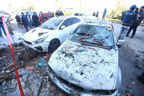 Mohammad Abushama / Anadolu via Getty Images Two damaged cars covered in debris are pictured with crowds of people on the street in the background with rubble all around, after an Israeli missile attack on the coastal road in Sidon on Wednesday.