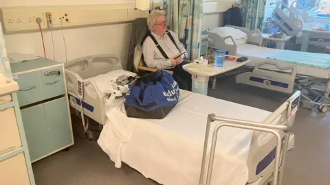 Patient Nick Wright pictured on the ward. He is sat between two beds. 