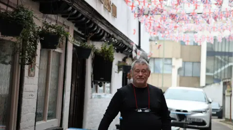 Steve Wood standing outside his pub. There's a lot of England flags hanging in the air behind him. He wears a black top and has glasses around his neck on a string