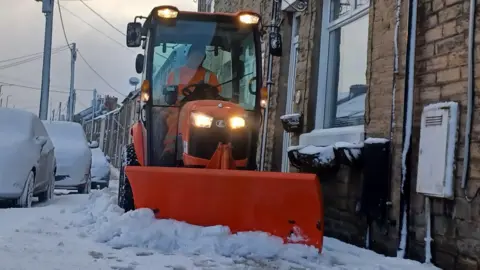 Durham County Council A man driving a bright orange gritter drives down a road covered in snow. Cars parked nearby are completely covered in snow. 