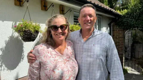 Zac Sherratt/BBC Steph and Neil Stephenson stood outside Fairacre in Haslemere. Neil has his arm around Steph, who is wearing a pink top and sunglasses. Neil is wearing a blue checked shirt. He has greying hair while Steph has blondish hair.
