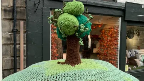 PA Media A crocheted tree made from brown and green wool sits on top of a post box with shops behind it
