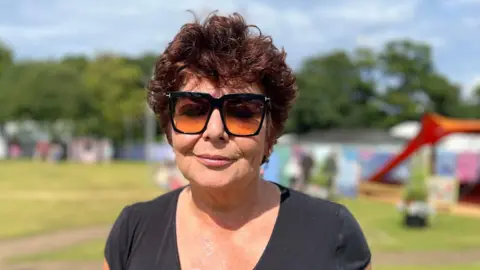 A woman with short dark red hair smiles at the camera. She wears a black top and dark sunglasses and stands in an outdoor setting with grass and trees behind her, as well as colourful constructions.