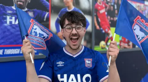Jack Richardson is holding two flags either side of him. He has a happy expression and has short brown hair. He is wearing black framed glasses and a blue Ipswich Town FC football T-shirt.
