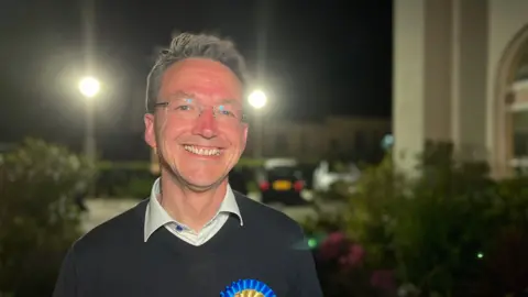 A picture of Kirsten Morel smiling at the camera. He has a black jumper and a white shirt below it, he is wearing a blue rosette.