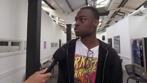 A young black man talking into a microphone in a college corridor. He is wearing a black hoodie and white t-shirt with an orange and red drawing